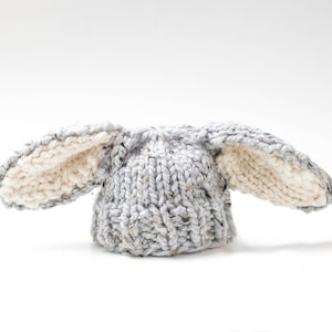 May include: A gray and white knitted hat with bunny ears. The hat is made of a soft, chunky yarn and has a slouchy fit.