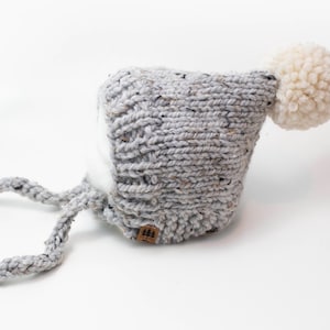 Könnte beinhalten: Eine graue gestrickte Baby-Mütze mit einem weißen Bommel oben und zwei langen Bändern. Die Mütze hat ein strukturiertes Strickmuster und ein kleines braunes Etikett mit einem Logo.