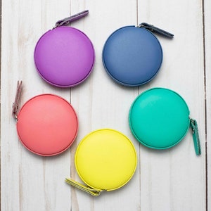 Puede incluir: Cinco cintas métricas de colores con diferentes estuches de colores: morado, azul, rosa, verde y amarillo. Las cintas métricas están dispuestas en círculo sobre una superficie de madera blanca.