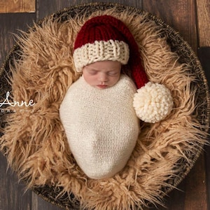 Op de afbeelding: Een pasgeboren baby gewikkeld in een witte gebreide cocon, met een rood-witte kerstmuts met een grote crèmekleurige pompon. De baby rust in een ronde, geweven mand gevoerd met pluizig, lichtbruin imitatiebont, tegen een donkere houten achtergrond.
