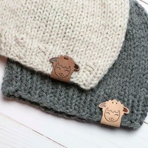 May include: Two knitted hats with a brown leather sheep applique. The hats are one grey and one cream.