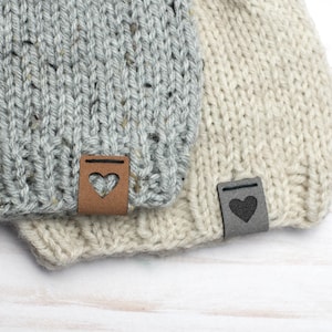 May include: Two knitted hats with leather heart-shaped tags. The hat on top is gray with a brown tag, and the bottom hat is beige with a black tag.