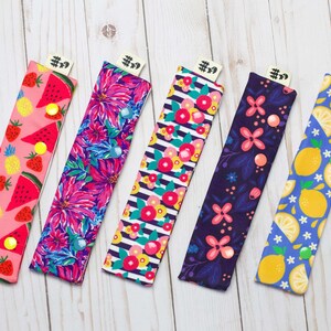 May include: Five colorful fabric covered pencil cases with different fruit patterns. The cases are pink, purple, blue, green, and yellow with watermelon, flowers, lemons, and berries. Each case has a black tag with the text "#9" on it.