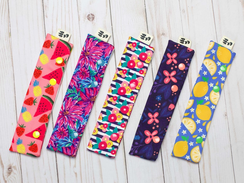 May include: Five colorful fabric covered pencil cases with different fruit patterns. The cases are pink, purple, blue, green, and yellow with watermelon, flowers, lemons, and berries. Each case has a black tag with the text "#9" on it.