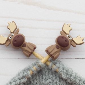 Könnte beinhalten: Zwei braune Elch-Stricknadelstopper mit hellbraunen Geweihen stecken in einem grauen Strickstoff. Die Stopper haben ein cartoonartiges Design. Der Hintergrund ist ein helles Holzmaserungsmuster.