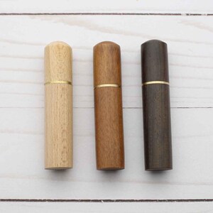 May include: Three cylindrical wooden cases arranged vertically, each featuring a smooth finish and a thin gold-toned metal band separating the cap from the base. The cases are presented in light, medium, and dark brown wood grain tones.