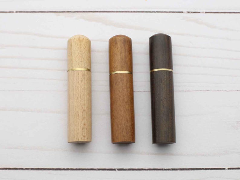 May include: Three cylindrical wooden cases arranged vertically, each featuring a smooth finish and a thin gold-toned metal band separating the cap from the base. The cases are presented in light, medium, and dark brown wood grain tones.