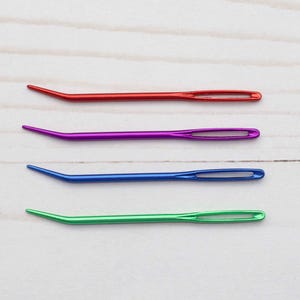May include: Four colorful plastic needle threaders, red, purple, blue, and green, are arranged in a row on a white wooden surface.
