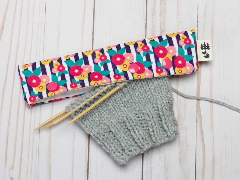 May include: A gray knitted fabric with wooden knitting needles and a floral patterned fabric needle holder. The needle holder has a small tag with a tree and fox design.