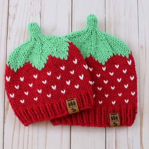 Op de afbeelding: Twee rood en groen gebreide aardbeienmutsen met witte zaadjes. Elke muts heeft een groene steel en een klein bruin label met een boomlogo. De mutsen zijn ontworpen om op aardbeien te lijken.