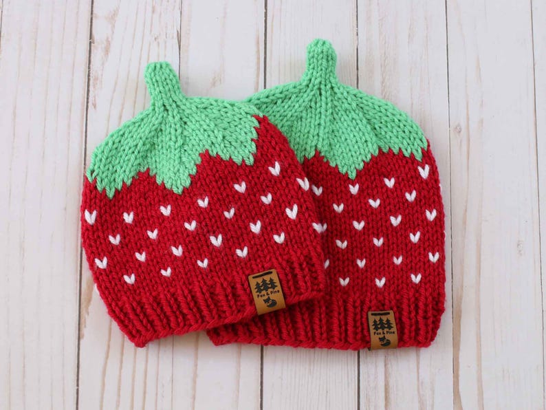 Puede incluir: Dos gorros de fresa tejidos en rojo y verde, con patrones de semillas blancas. Cada gorro tiene un tallo verde y una peque&ntilde;a etiqueta marr&oacute;n con un logotipo de &aacute;rbol. Los gorros est&aacute;n dise&ntilde;ados para parecerse a fresas.