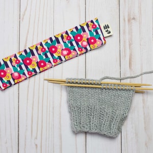 May include: A gray knitted fabric with wooden knitting needles, a floral patterned fabric needle case with a tag that says "#Knit", and a white wooden background.