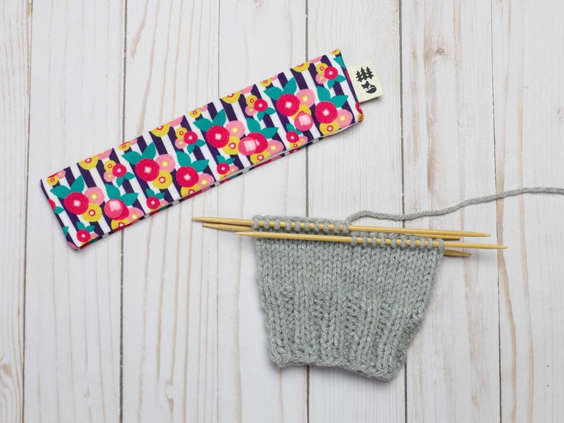 May include: A gray knitted fabric with wooden knitting needles, a floral patterned fabric needle case with a tag that says "#Knit", and a white wooden background.