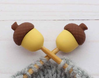 May include: Two acorn-shaped stitch markers with brown caps and yellow bodies are crossed over a gray knitted project. The stitch markers are on wooden sticks, and the background is a white wooden surface.