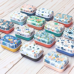 May include: A collection of 14 small, rectangular tin boxes with various floral patterns. The boxes are arranged in rows on a light wood surface. Each box has a different floral design, including blue, pink, green, and white flowers.