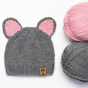 May include: A gray knitted hat with pink knit ears and a brown leather tag that says "Fox & Fox". Two balls of yarn, one pink and one gray, are also in the image.