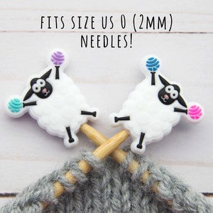 Può includere: Due ferri da maglia in legno con marcatori di punto a forma di pecora bianca. Le pecore tengono gomitoli di lana blu, rosa e viola. I ferri sono incrociati e il testo "FITS SIZE US 0 (2MM) NEEDLES!" è sopra i ferri.