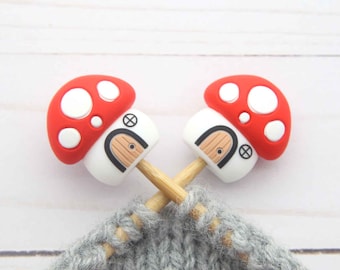 May include: Two mushroom-shaped stitch markers with red caps and white spots, white bases with a door design, and wooden stems. The markers are on knitting needles, with gray yarn in the foreground.