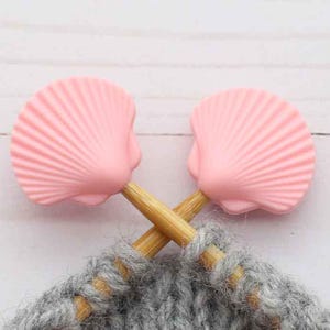 Puede incluir: Un par de agujas de tejer de madera con topes en forma de concha rosa, cruzadas sobre una sección de tejido de punto gris. Las agujas son de madera marrón claro y las conchas son rosas.