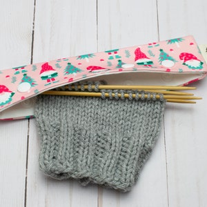 May include: A pink knitting needle case with a festive gnome and Christmas tree pattern. The case holds wooden knitting needles and a partially knitted gray sleeve. A white snap closure secures the case.
