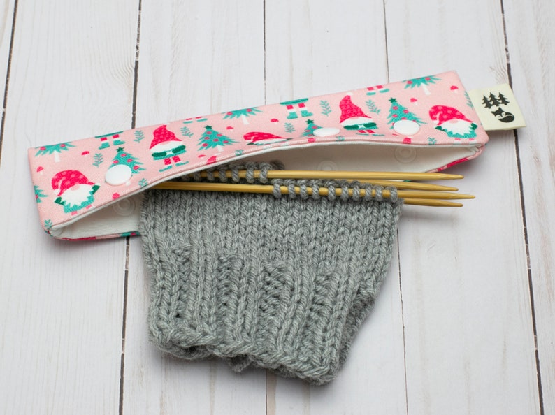 May include: A pink knitting needle case with a festive gnome and Christmas tree pattern. The case holds wooden knitting needles and a partially knitted gray sleeve. A white snap closure secures the case.