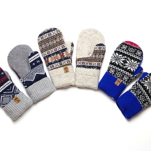 Op de afbeelding: Vijf paar gebreide wanten met verschillende patronen. De wanten zijn in verschillende kleuren, waaronder rood, blauw, zwart, wit en grijs. Elke want heeft een klein bruin label met het woord "Tree" erop.