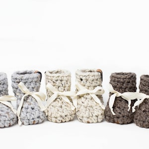 May include: Six handmade crochet baby booties in various neutral colors: gray, off-white, and brown. Each bootie has a cream-colored ribbon tied in a bow and a small leather tag. Perfect for a baby shower gift.