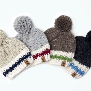 May include: Four knitted beanies with pom-poms in different colors, each with a white band and a colored stripe. The beanies have a brown leather patch with a tree logo.