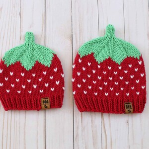 Puede incluir: Dos gorros de fresa tejidos. Los gorros son rojos con detalles blancos en forma de coraz&oacute;n y copas verdes con forma de hoja. Cada gorro tiene una peque&ntilde;a etiqueta marr&oacute;n con un logotipo de &aacute;rbol.