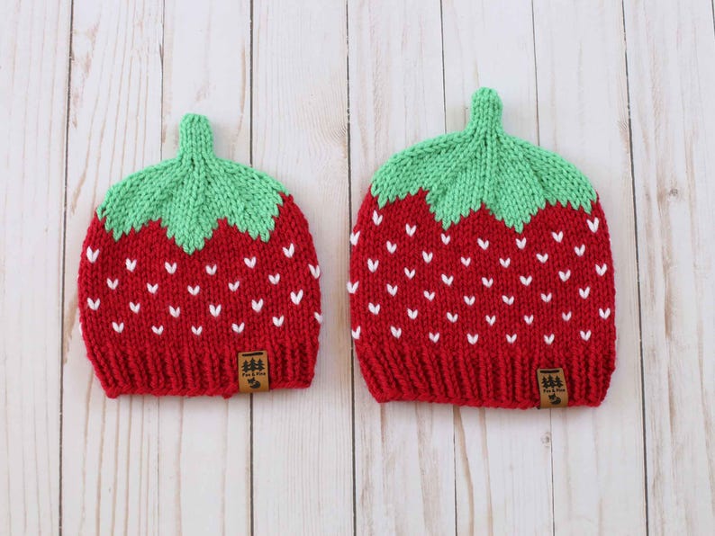 Puede incluir: Dos gorros de fresa tejidos. Los gorros son rojos con detalles blancos en forma de coraz&oacute;n y copas verdes con forma de hoja. Cada gorro tiene una peque&ntilde;a etiqueta marr&oacute;n con un logotipo de &aacute;rbol.