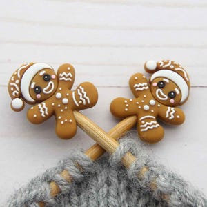 May include: Two gingerbread man stitch markers with white icing hats and details, resting on wooden knitting needles. The markers are brown with white accents and are positioned over a grey knitted fabric.