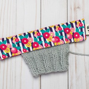 May include: A gray knitted hat with a floral fabric band. The band has a white background with black stripes and pink, yellow, and green flowers. The band has two pink snap buttons. A small tag with a black silhouette of a fox and trees is attached to the band.