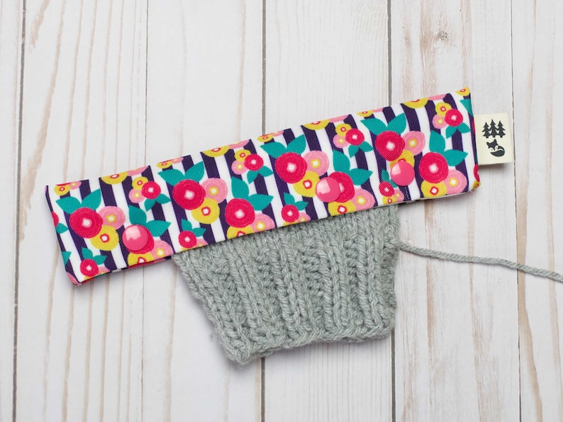 May include: A gray knitted hat with a floral fabric band. The band has a white background with black stripes and pink, yellow, and green flowers. The band has two pink snap buttons. A small tag with a black silhouette of a fox and trees is attached to the band.