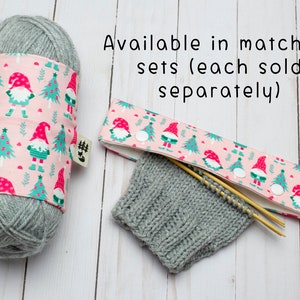 May include: A gray yarn ball wrapped with a pink fabric featuring a gnome and Christmas tree pattern. A matching knitting needle case and knitted gray swatch are also shown. Text reads: Available in matching sets (each sold separately).