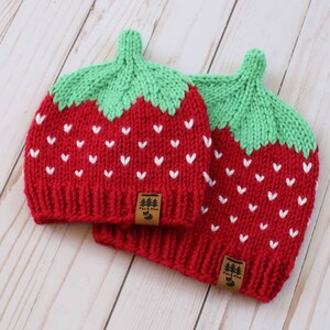 Puede incluir: Dos gorros rojos de punto con la parte superior verde, que parecen fresas. Cada gorro tiene detalles blancos en forma de coraz&oacute;n y una peque&ntilde;a etiqueta marr&oacute;n. Los gorros est&aacute;n sobre una superficie de madera clara.
