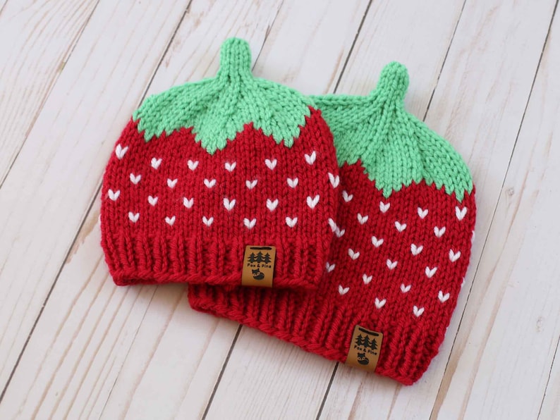 Puede incluir: Dos gorros rojos de punto con la parte superior verde, que parecen fresas. Cada gorro tiene detalles blancos en forma de coraz&oacute;n y una peque&ntilde;a etiqueta marr&oacute;n. Los gorros est&aacute;n sobre una superficie de madera clara.