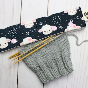 May include: A gray knitted fabric with two wooden knitting needles and a black and white fabric headband with a sheep pattern and pink buttons. The headband has a small tag that says "Fox & Pine".