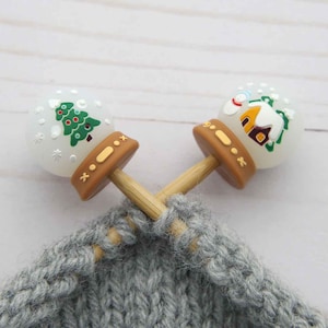May include: Two decorative snow globe stitch markers with brown bases and wooden sticks are inserted into a grey knitted fabric. One globe features a green Christmas tree, the other a house. The background is a white wooden surface.