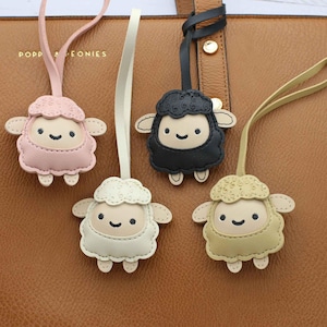 May include: Four sheep-shaped bag charms with smooth faces and textured wool tops, each featuring a strap for attachment. The charms are coloured in light pink, cream, black, and beige, and they are displayed against a brown textured bag.