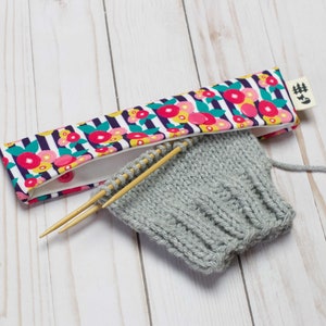 May include: A gray knitted hat with wooden knitting needles and a floral patterned fabric needle holder with the text "#5".