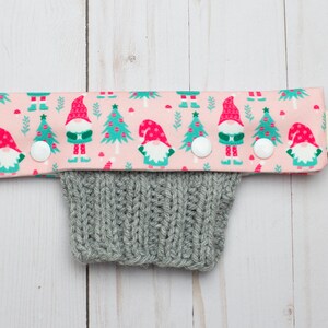 May include: A pink fabric cuff with a repeating pattern of gnomes and Christmas trees, attached to a gray knitted sleeve. The cuff has white buttons and a small tag with a forest and fox logo.