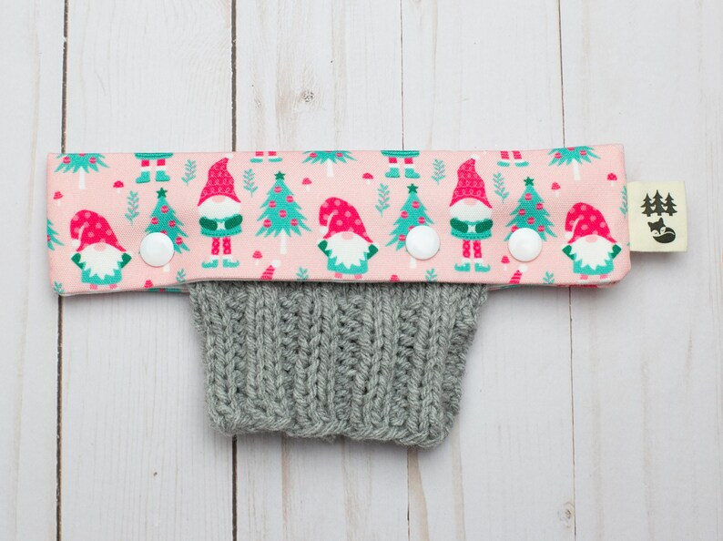 May include: A pink fabric cuff with a repeating pattern of gnomes and Christmas trees, attached to a gray knitted sleeve. The cuff has white buttons and a small tag with a forest and fox logo.