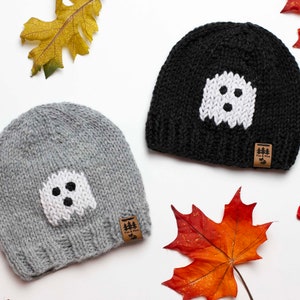 May include: Two knitted hats with ghost designs. The hats are grey and black with white ghost faces. The hats have brown leather tags with the text "Fox & Pine" on them.