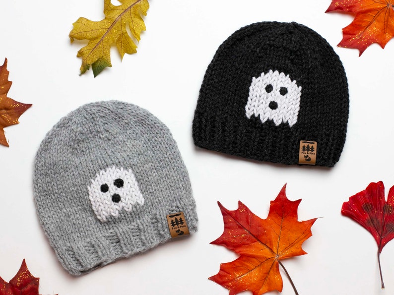 May include: Two knitted hats with ghost designs. The hats are grey and black with white ghost faces. The hats have brown leather tags with the text "Fox & Pine" on them.