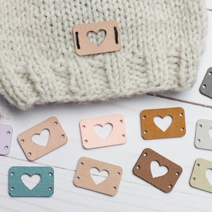May include: Assortment of ten heart-shaped leather labels in various colours, including pink, brown, beige, grey, and teal. Each label has a heart-shaped cutout and four holes for sewing.