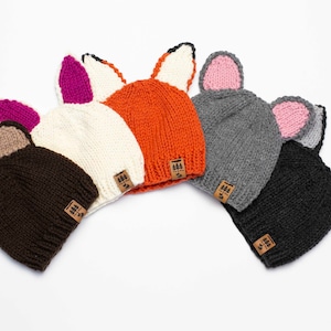 May include: A collection of six knitted beanies in various colors, each featuring animal ears. The hats include brown bear ears, white bunny ears, orange fox ears, gray cat ears, and black cat ears. Each hat has a small, rectangular tag.