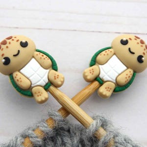 Peut inclure: Deux marqueurs de point en forme de tortue avec des visages souriants, des taches marron et des carapaces vertes sont fixés à des aiguilles à tricoter en bois. Les aiguilles sont croisées sur de la laine grise, suggérant un projet de tricot. Les marqueurs ajoutent une touche fantaisiste.