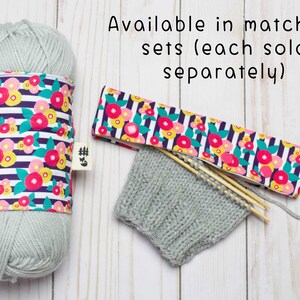 May include: A set of matching knitting accessories in a floral print. The set includes a yarn bowl, a needle case, and a knitted hat. The yarn bowl and needle case are both made of fabric with a white background and a pattern of pink, yellow, and green flowers on black stripes. The hat is a light gray color and is made of knitted yarn. The text "Available in matching sets (each sold separately)" is visible in the image.