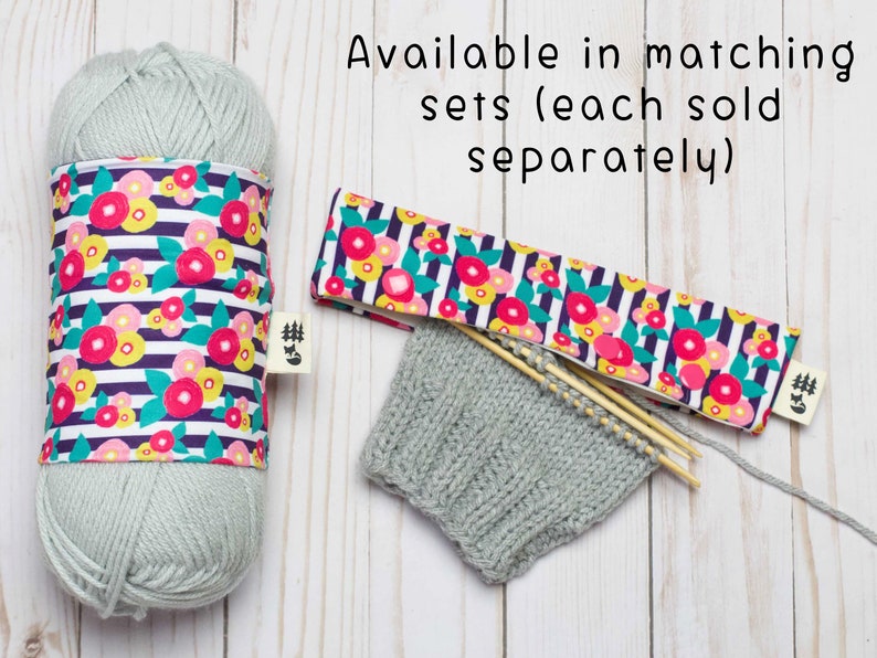 May include: A set of matching knitting accessories in a floral print. The set includes a yarn bowl, a needle case, and a knitted hat. The yarn bowl and needle case are both made of fabric with a white background and a pattern of pink, yellow, and green flowers on black stripes. The hat is a light gray color and is made of knitted yarn. The text "Available in matching sets (each sold separately)" is visible in the image.