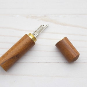 May include: A cylindrical wooden needle case with a brass screw-top lid, shown open to reveal several silver-toned metal sewing needles stored inside. The case features a smooth, polished brown wood grain finish and a metallic brass threaded neck.